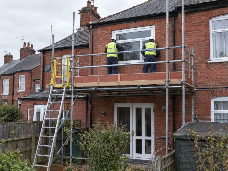 Builders on a guarded scaffold fitting a window at the rear of a terraced house